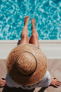 woman sitting on poolside setting both of her feet on pool - summer skincare