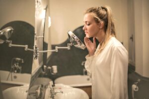 Side view of blond woman in white blouse standing in bathroom looking closely at face skin in small mirror