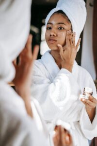 Young black female in white robe and towel on head applying moisturizing cream on face while standing in bathroom | Morning vs Night Skincare Routines