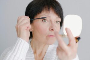 A senior woman in a cozy sweater applying mascara with a focused expression indoors.