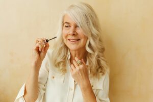 Elderly woman with white hair smiling while applying makeup indoors.