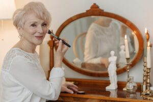 Senior woman applying makeup with brush by a vintage mirror, showcasing elegance and grace.