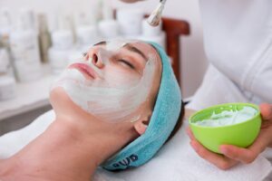 woman lying on blue towel with white cream on face | Facial Treatment