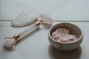 Close-up of a skincare setup featuring a gua sha tool, jade roller, and pink cream in a bowl.