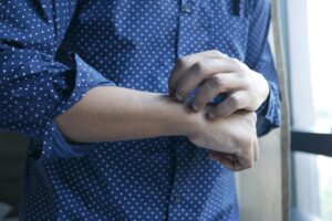 A man in a polka dot shirt scratching his arm next to a window, symbolizing discomfort or irritation.