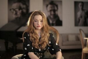 A woman with long wavy hair, seated indoors, displaying a pensive expression against a blurred background. | Difficult Salon Clients