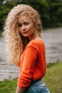 Young woman with curly hair poses by a riverside in an orange sweater, showcasing fashion and beauty.