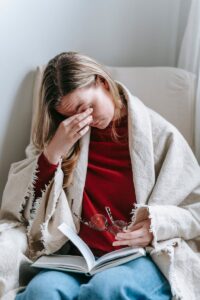 Woman sitting in an armchair with a blanket, holding a book and rubbing her eyes, wearing a red sweater.