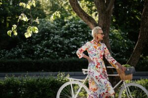 Stylish senior woman with a bicycle and floral dress enjoying nature outdoors. | Mature skin