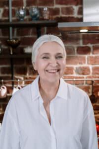 Senior woman smiling warmly in a cozy brick kitchen with rustic decor.